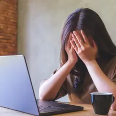 Kobieta z dłońmi na twarzy, przy laptopie, z kubkiem kawy. Objawy wypalenia zawodowego, czas na L4.