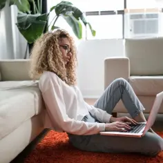 Kobieta z kręconymi włosami pracuje na laptopie na dywanie, opierając się o kanapę. Czy na macierzyńskim można pracować? Tak, zdalnie.