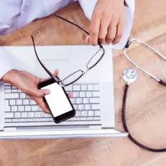 Lekarz z okularami i telefonem nad laptopem, obok stetoskop. Dostęp do informacji o dobrowolnym ubezpieczeniu zdrowotnym.