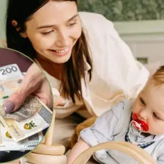 Mama z uśmiechem trzyma banknoty 200 zł, symbolizujące 1000 zł na drugie dziecko. Obok bawi się maluch.