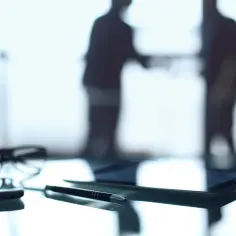 Okulary, długopis i tablet na biurku. W tle sylwetki dwóch mężczyzn, być może omawiających kwestię opłaty skarbowej.