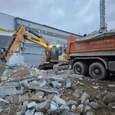 Żółty koparka pracuje przy rozbiórce gruzowiska. Obok stoi pomarańczowa wywrotka, gotowa zabrać urobek.