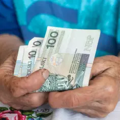 Dłonie starszej osoby trzymają banknoty 100 zł. To symbol oszczędności na ubezpieczenie emerytalne.