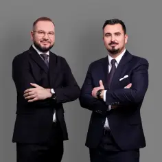 Dwóch mężczyzn w garniturach stoi ze skrzyżowanymi rękami na szarym tle.