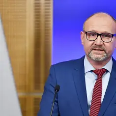 Prokurator krajowy w garniturze i okularach przemawia przy mikrofonie, obok polskiej flagi.