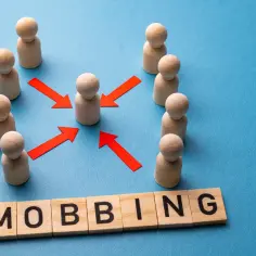 Drewniane figurki ludzi otaczają jedną, a czerwone strzałki wskazują na nią. Słowo "MOBBING" ułożone z klocków symbolizuje problem, który reguluje kodeks pracy.