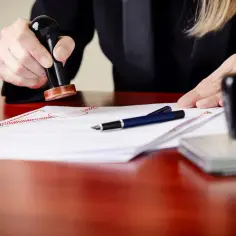 Dłoń notariusza stempluje dokument. Obok leży długopis i pieczęć.