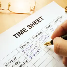 Dłoń pisze na karcie czasu pracy, rejestrując godziny. Okulary i długopis leżą obok.