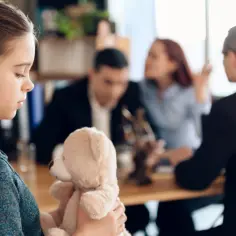 Smutna dziewczynka z misiem w ramionach, w tle kłócący się rodzice i kurator sądowy.