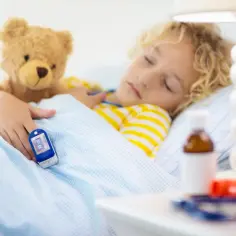 Chory chłopiec śpi z pluszowym misiem, a na palcu ma pulsoksymetr. Obok leki, wszystko dla l4 na dziecko.