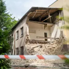 Widoczny fragment budynku po zawaleniu się części ściany i dachu. Gruz i elementy konstrukcyjne leżą na ziemi. Wstęga ostrzegawcza odgradza teren.