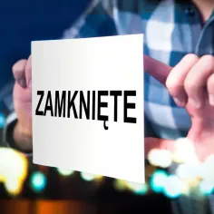 Ręka przykleja kartkę z napisem "ZAMKNIĘTE" na szybie, sygnalizując zamknięcie działalności gospodarczej.
