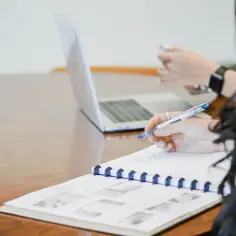 Ręka pisze notatki, przygotowując podanie o przedłużenie umowy o pracę uzasadnienie. Obok laptop i telefon.