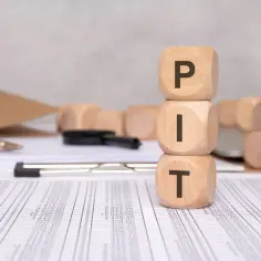 Drewniane kostki z literami PIT ułożone w stos. Dokumenty i długopis sugerują rozliczenie podatkowe, być może ulga rehabilitacyjna 2026.