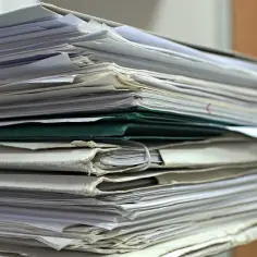Stos dokumentów, wśród nich zielona teczka, sugeruje archiwum lub biuro z ważnymi sprawami, jak lista płac.