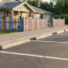 Puste miejsca parkingowe z wyznaczonymi liniami i ogranicznikami. W tle kolorowy płotek i budynek. Wymiary miejsca parkingowego są standardowe.