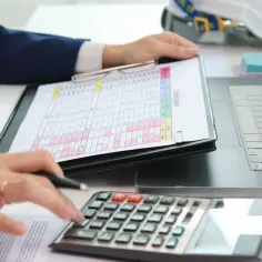 Osoba analizuje raport kosztów, używając kalkulatora i laptopa. W tle kask budowlany.