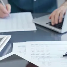 Osoba pracuje przy laptopie, analizując dokumenty. Obok leży kalkulator i okulary. To idealne miejsce do podpisania umowy zlecenie do stażu pracy.