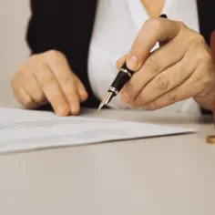 Dłoń trzymająca pióro nad dokumentem, obok pieczęć.
