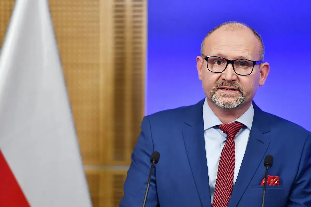 Prokurator krajowy w niebieskim garniturze, z czerwoną poszetką i krawatem, przemawia przy mikrofonie. Obok widoczna polska flaga.