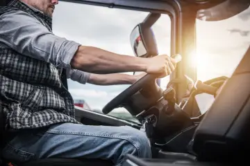 Mężczyzna w kratę i jeansach prowadzi ciężarówkę. Jego dłonie spoczywają na kierownicy, a kluczyki wiszą przy stacyjce. To jego świadectwo kierowcy.