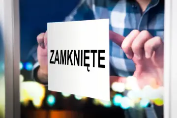 Ręka przykleja kartkę z napisem "ZAMKNIĘTE" na szybie, sygnalizując zamknięcie działalności gospodarczej.